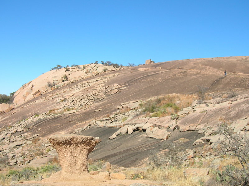 Rock pedestal with exfoliating slabs in the background. Enchanted Rock, Texas. This granite is dated at 1.1 billion years (1.1Ga), likely the product of tectonic convergence between Laurentia (North America)and Gonawaland (South America+Africa), resulting in the super continent, Rondinia. The rock pedestal is the result of differential erosion: the base experiences greater chemical weathering than the upper part due to standing water at the base. Case-hardening on the surface of the top of the pedestal helps resist weathering. Exfoliation is explained as the reaction to the removal of overlying pressure (that is, rocks removed by erosion). Rocks exposed to such removal of overburden experience expansion: the result is exfoliation sheets. At Enchanted Rocks, one can see the exfoliated sheets sliding downslope.<br>