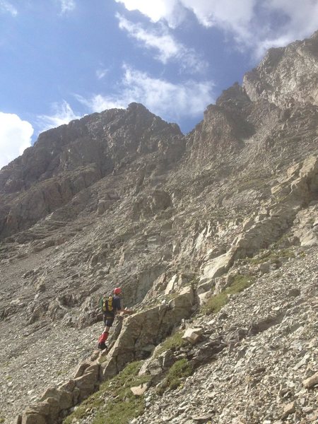 The middle section of the route, the "vegetated shelf" heading towards the red scree shoulder (which is in the notch on the skyline above the climber) on the North ridge. Summit highpoint on left.