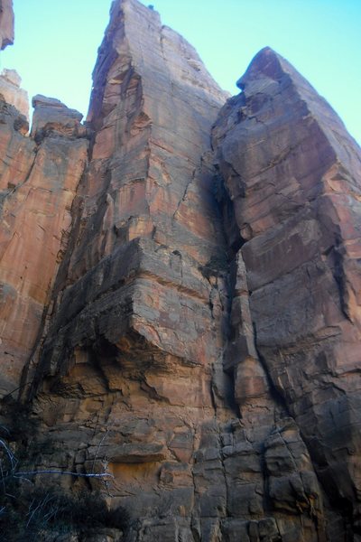 View from the jungle. Route starts at the veg ledge, swoops up and right to gain the tree ledge. Belay move left to arête. Obvious ramp leads up right. Cracks on the main face to the top.