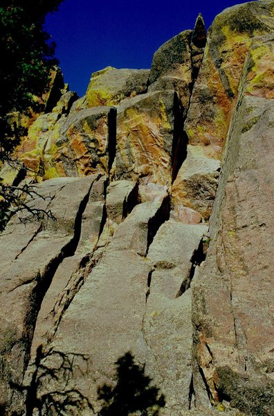 The route begins just right of the tree shadow and trends rightish into a short, right-facing dihedral and a wide inset, to the overhanging crack, which is followed up to and over the protruding wedged block near the top. This photo was taken on 10/15/83.