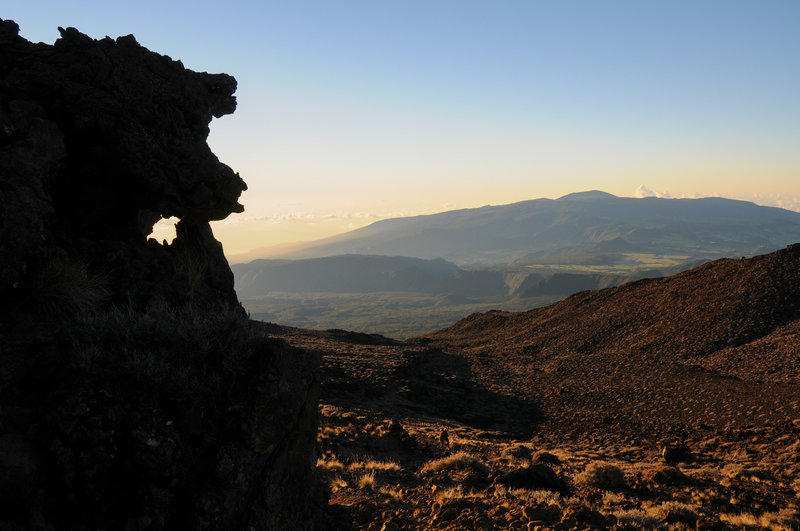 Piton de la Fournaise in the distant early morning light as a hiker descends the summit of Piton des Neiges near cool volcanic rock formations.
